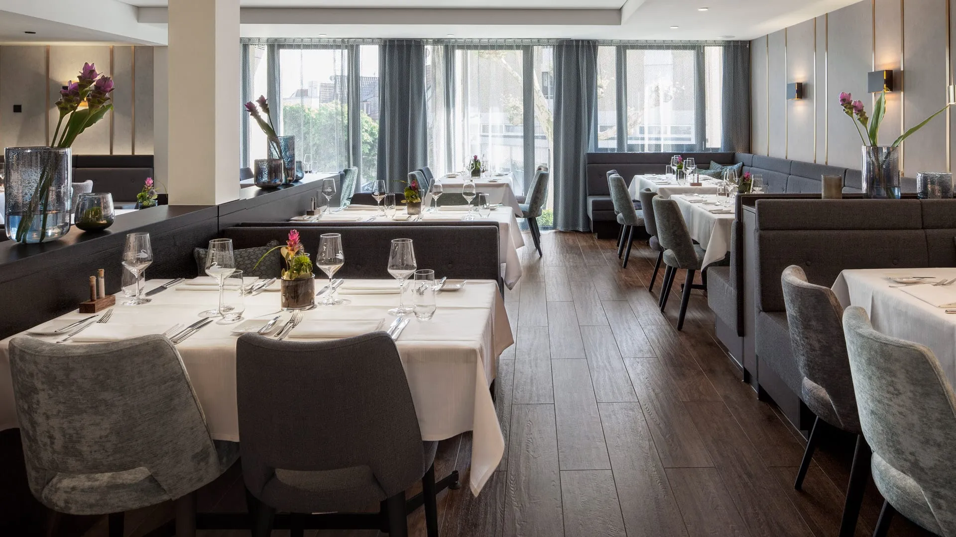 Schickes Restaurant mit gedeckten Tischen, grauen Stühlen und Blumenvasen, Holzboden, große Fenster mit Vorhängen.
