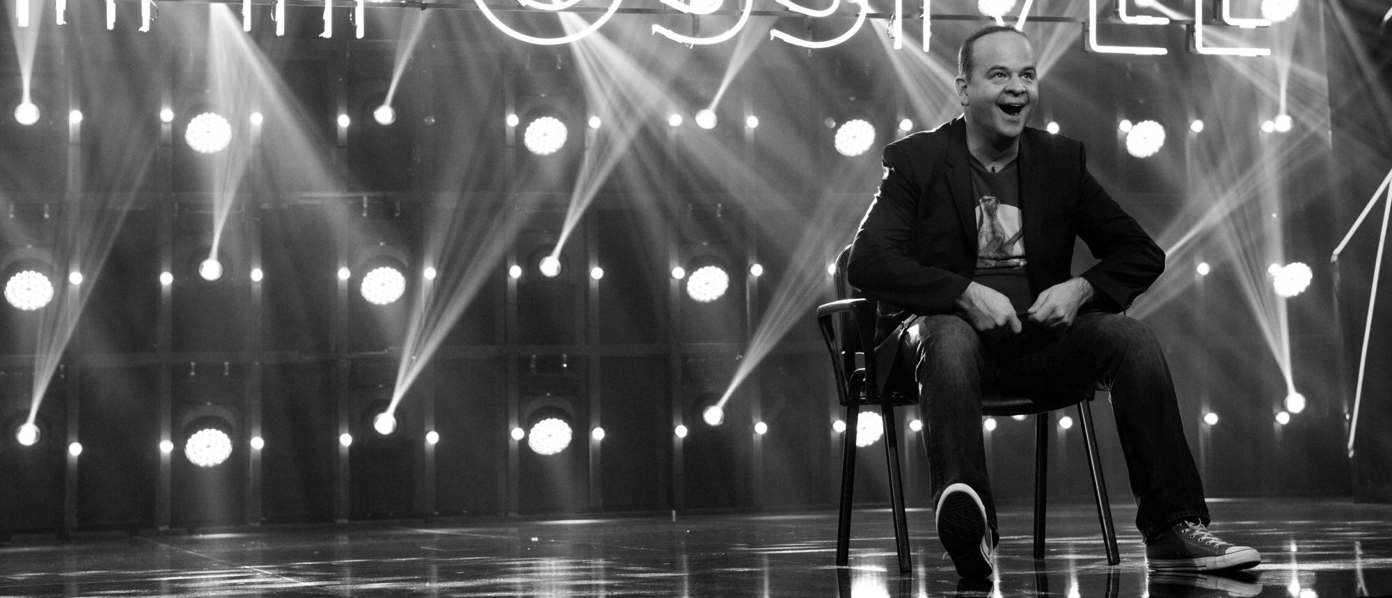 Mann sitzt lachend auf Bühne, umgeben von Scheinwerfern und Nebel, trägt Blazer und Jeans. Stimmungsvolles Licht.