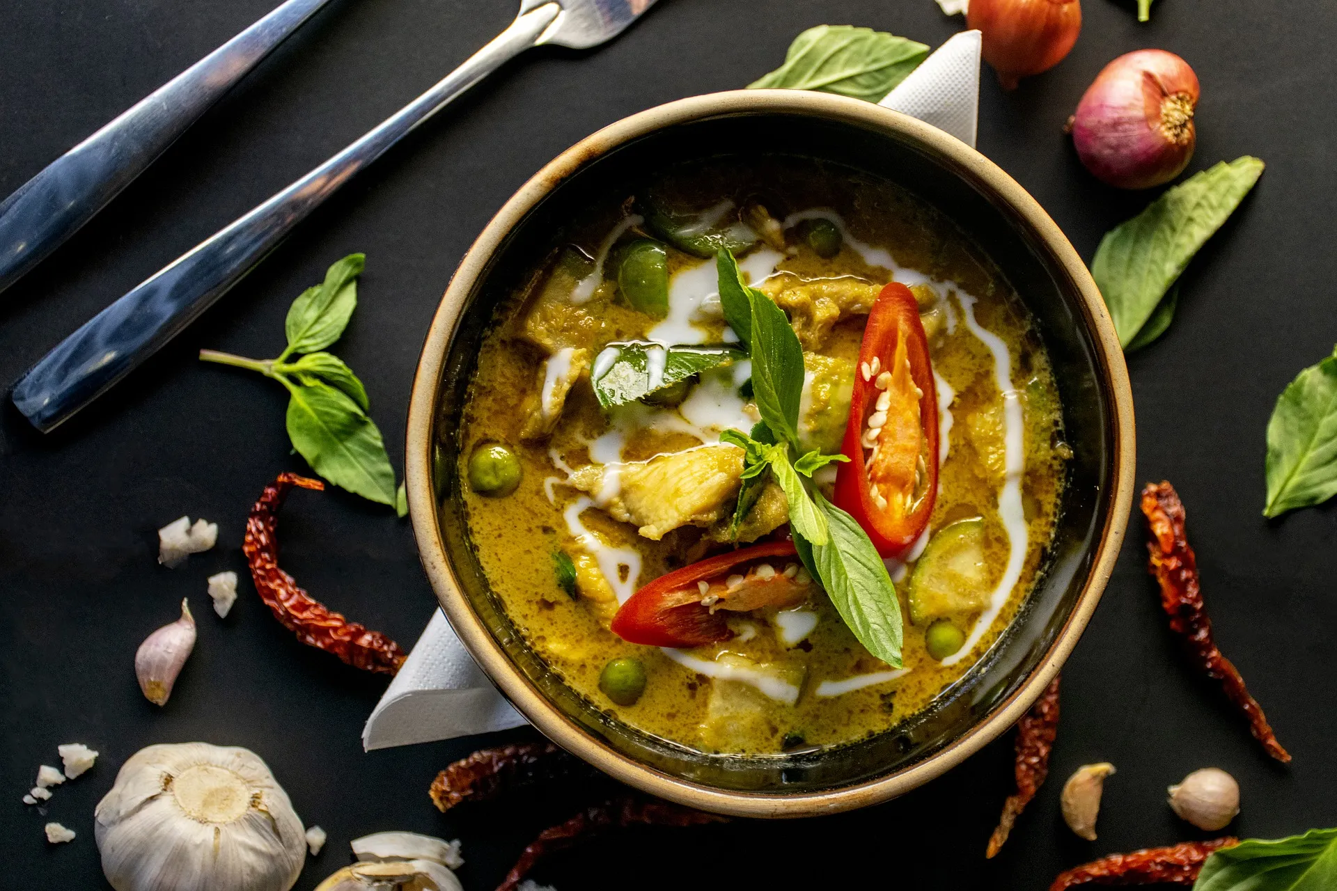 Grüne Curry-Suppe in Schale, garniert mit roter Chili und Basilikumblättern, umgeben von Knoblauch, Chilis und Besteck auf schwarzem Tisch.