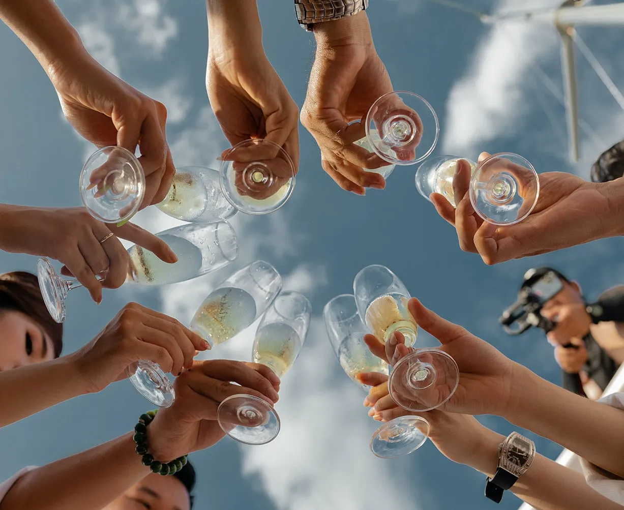 Mehrere Personen stoßen mit Sektgläsern an, von unten fotografiert, Himmel im Hintergrund.