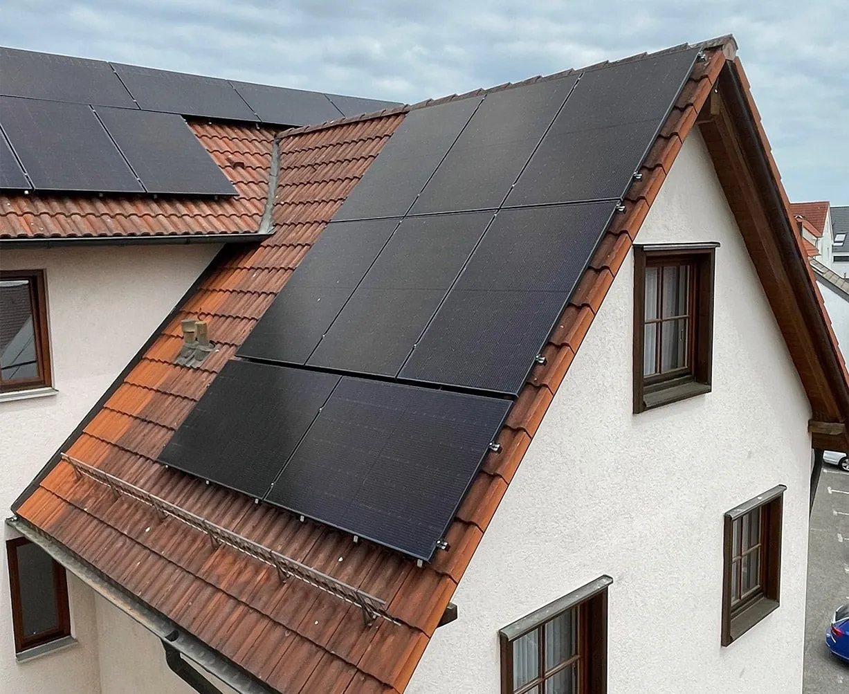 Solaranlagen auf dem Dach eines Hauses, grauer Himmel im Hintergrund, mehrere Fenster sichtbar.