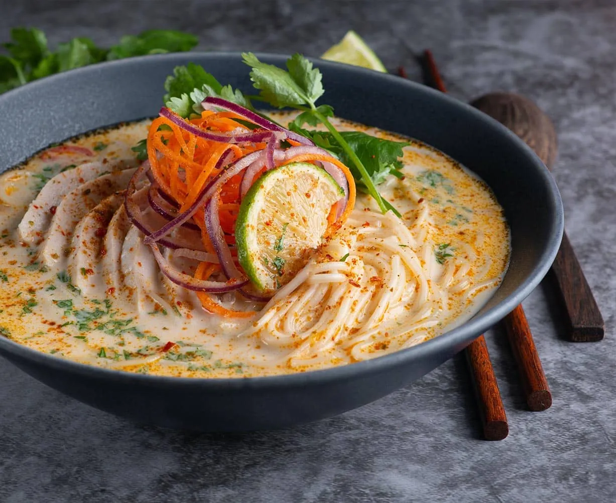 Schale mit cremiger Nudelsuppe, garniert mit Limonenscheibe, roten Zwiebeln, Karottenstreifen und Korianderblättern.
