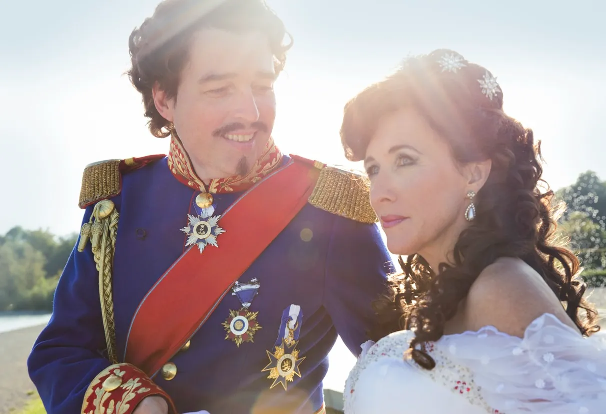 Ein Mann in einer blau-roten Uniform mit Auszeichnungen und eine Frau im weißen Kleid stehen draußen, Sonne im Hintergrund.