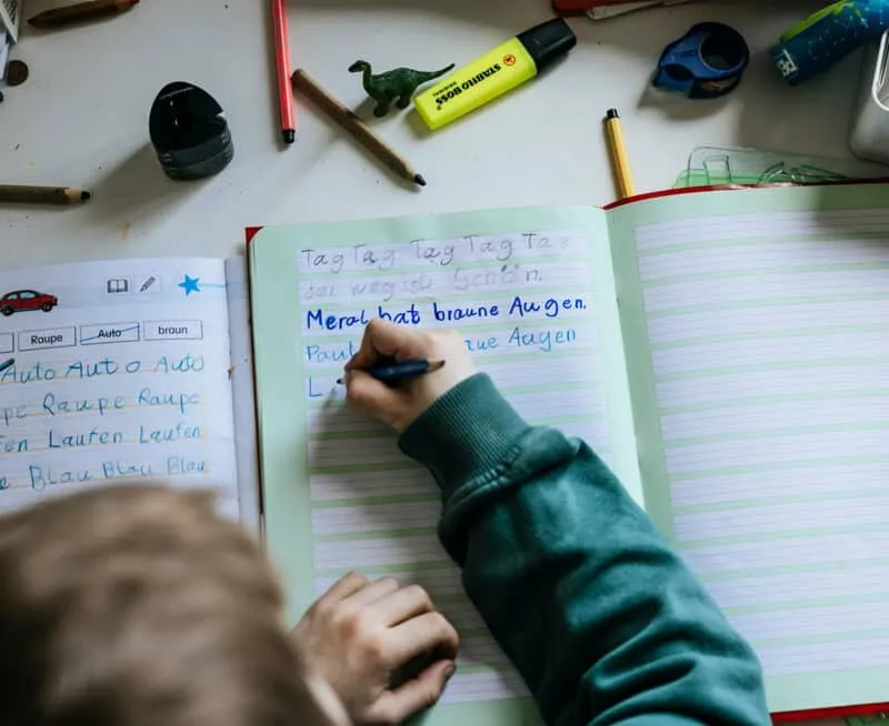 Ein Kind schreibt mit einem Stift in ein Heft, umgeben von Stiften und Schreibwaren auf einem Tisch.