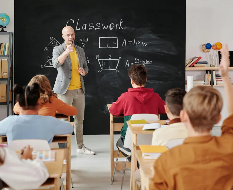 Ein Lehrer erklärt Geometrie an der Tafel, während Schüler in einem Klassenzimmer aufpassen und sich melden.