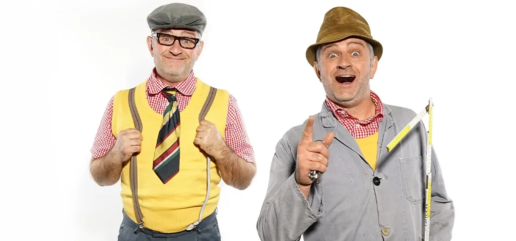 Zwei Männer in bunter Kleidung und Hüten, einer mit Krawatte, der andere mit Maßstab, vor weißem Hintergrund, beide lächelnd.