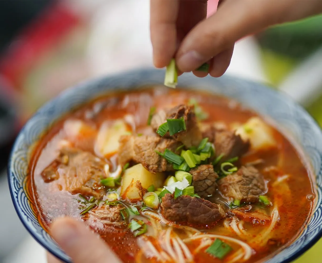 Hand streut Frühlingszwiebeln über eine Schale mit Nudelsuppe, Rindfleischstücken und Brühe.