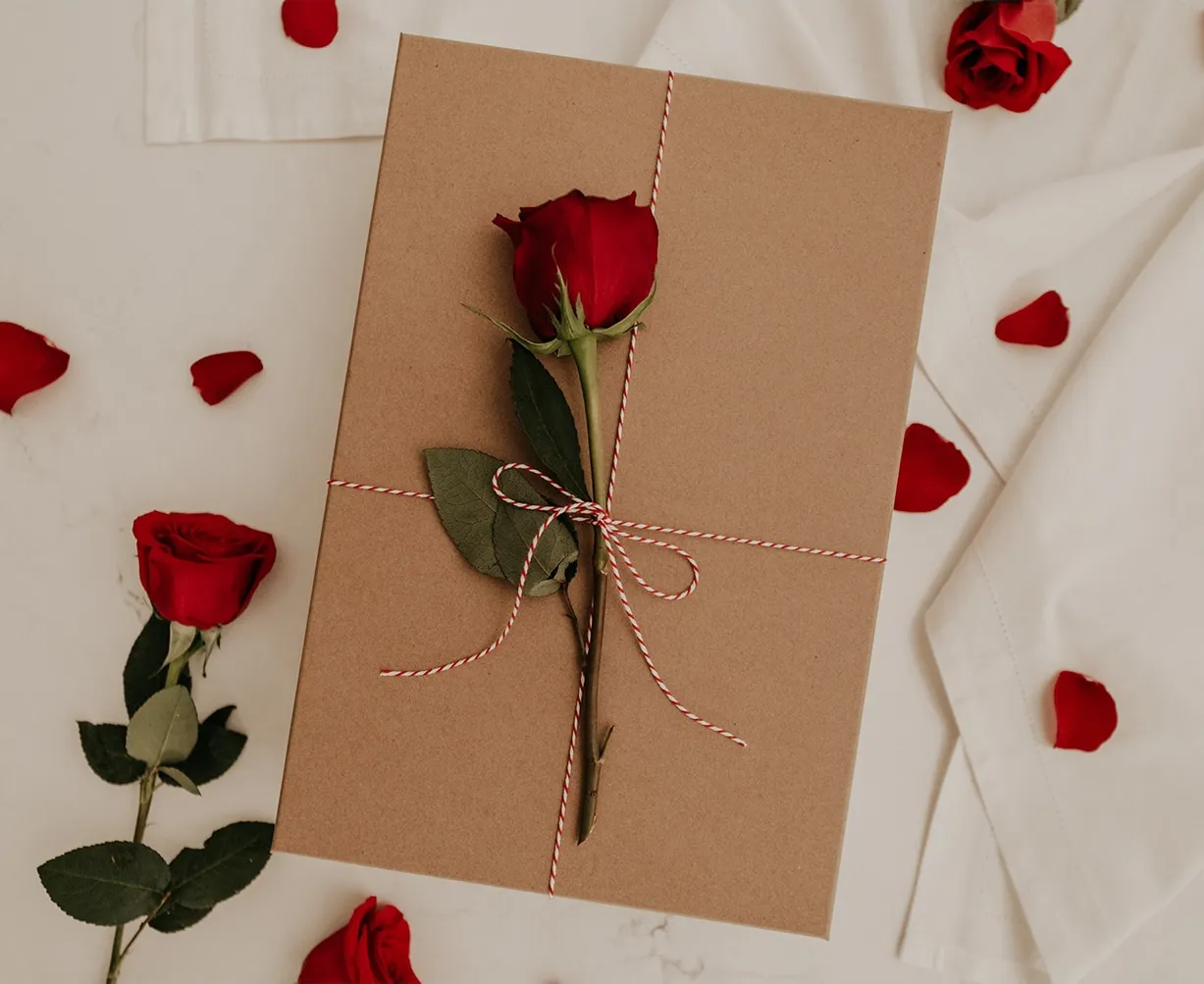 Braune Schachtel mit roter Rose, umwickelt mit Kordel, auf weißem Untergrund, umgeben von Rosenblättern und weiteren Rosen.
