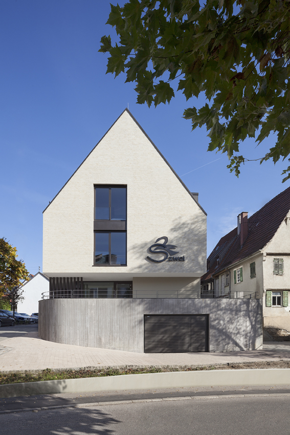 Modernes Gebäude mit Schrägdach und großem Fenster, moderner Schriftzug S auf der Vorderseite, blauer Himmel, Bäume.