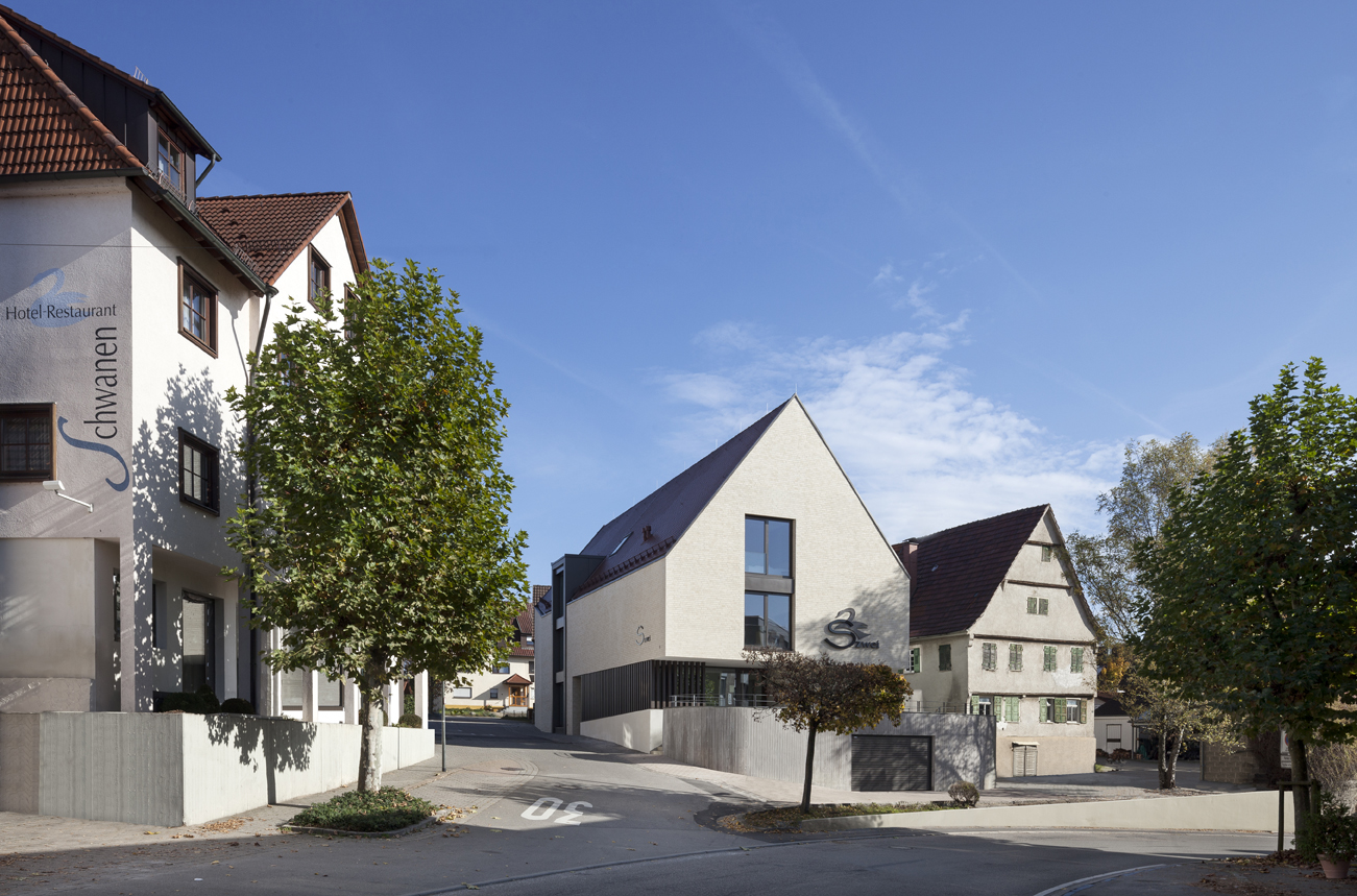 Modernes Gebäude mit spitzem Dach neben traditionellem Fachwerkhaus; klarer blauer Himmel, Bäume im Vordergrund.
