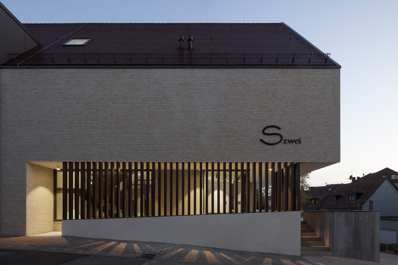 AQ460-13 Moderne Gebäudeaußenansicht bei Dämmerung, horizontale Holzlatten und beleuchteter Eingang, Schriftzug S zwei an der Fassade.