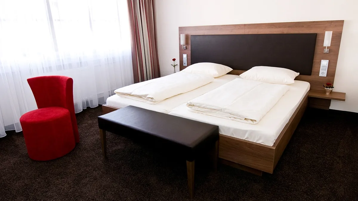 Doppelzimmer-Superior Doppelbett mit weißer Bettwäsche, schwarzer Bank, rotem Stuhl und Fenstervorhängen in einem Hotelzimmer.
