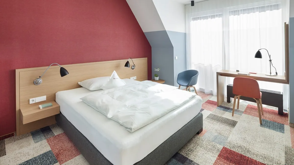 Modernes Hotelzimmer mit Doppelbett, Schreibtisch, zwei Stühlen, roter Wand und Licht von einem großen Fenster.