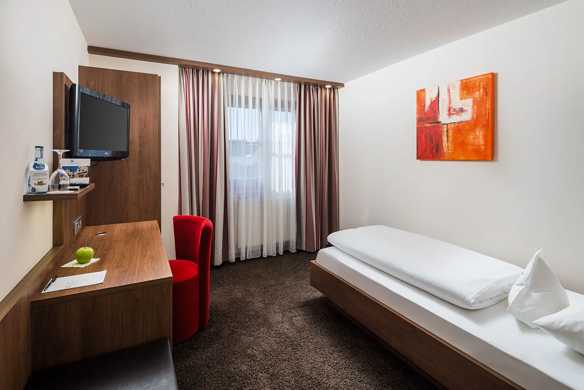 Hotel-Restaurant-Schwanen-Businesszimmer-01 Einzelzimmer mit Einzelbett, Schreibtisch, rotem Stuhl, Wand-TV und orangefarbenem Kunstwerk, modern eingerichtet.