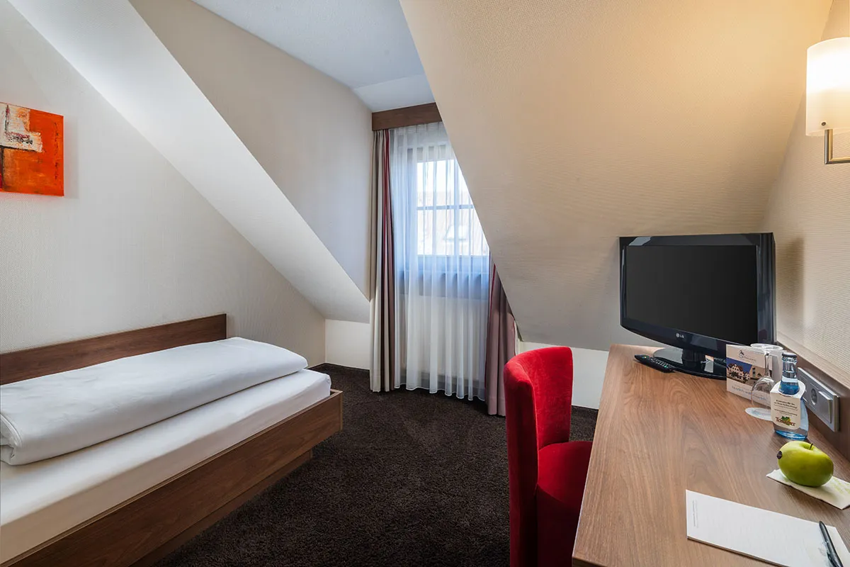 Hotel-Restaurant-Schwanen-Gaubenzimmer Kleines Zimmer mit Einzelbett, Schreibtisch, rotem Stuhl, Fernseher und schräger Decke. Fenster mit Vorhang und Tageslicht.
