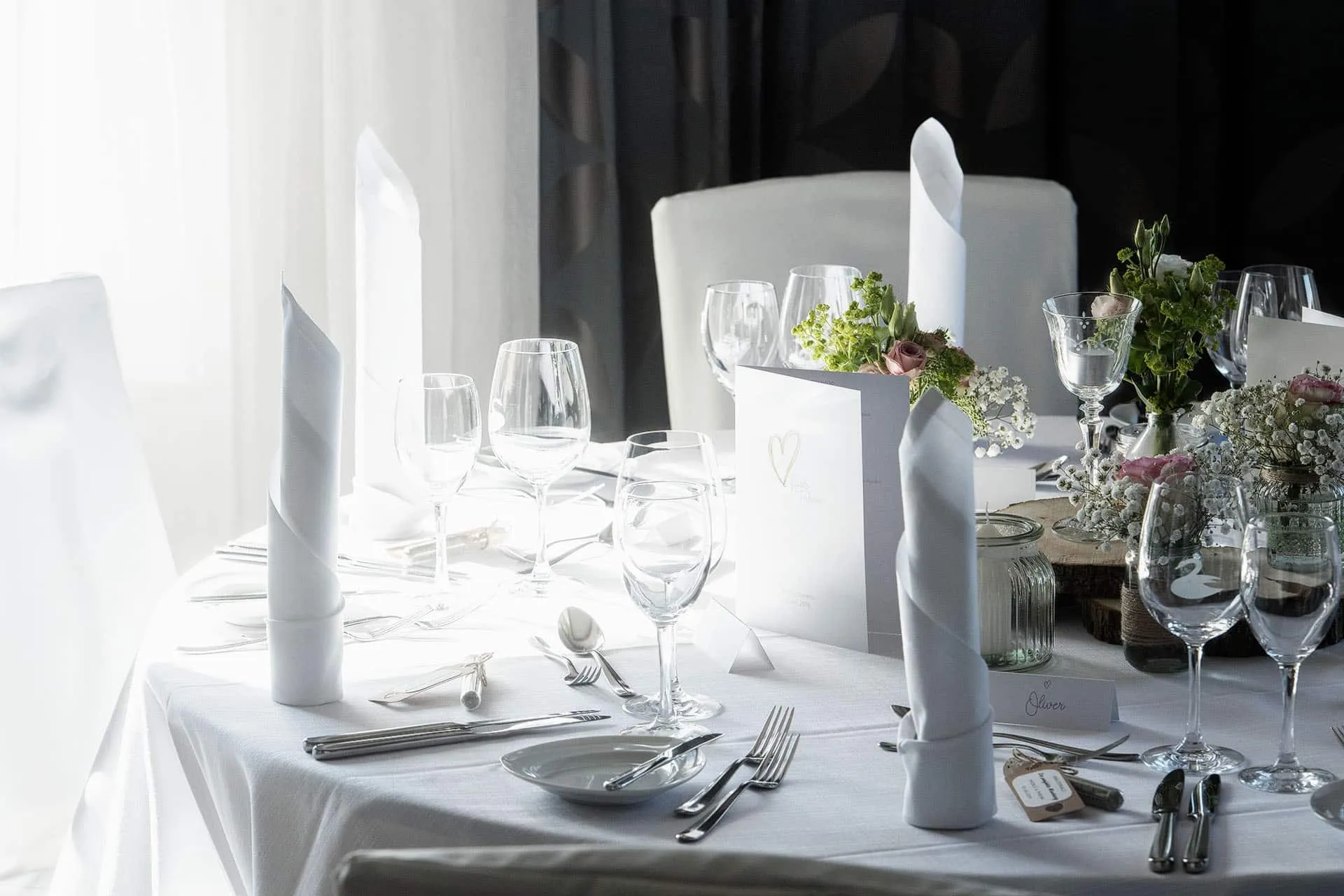 Elegant gedeckter Tisch mit weißen Servietten, Weingläsern und Blumenarrangements in heller, sonnendurchfluteter Umgebung.
