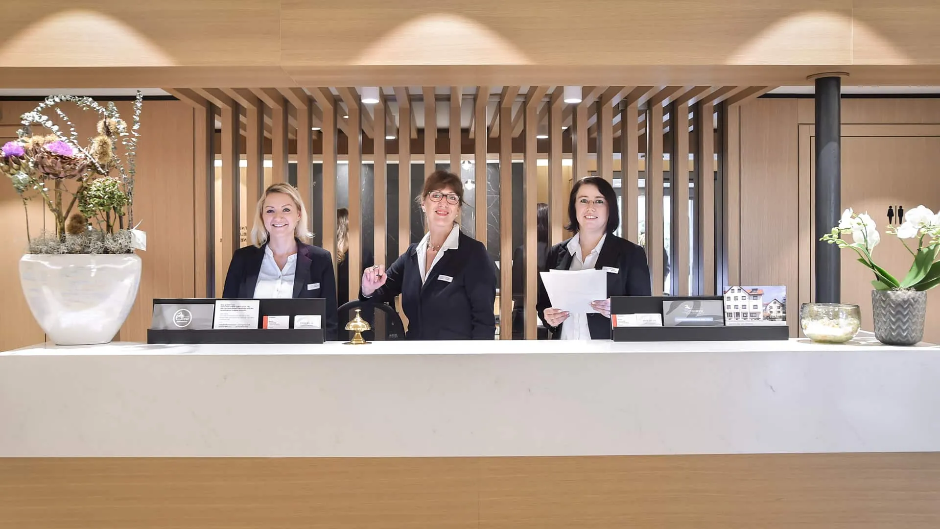 Drei Hotelmitarbeiter in Uniform stehen hinter einer Rezeption, lächelnd unter Holzdekor, mit Blumenarrangements.