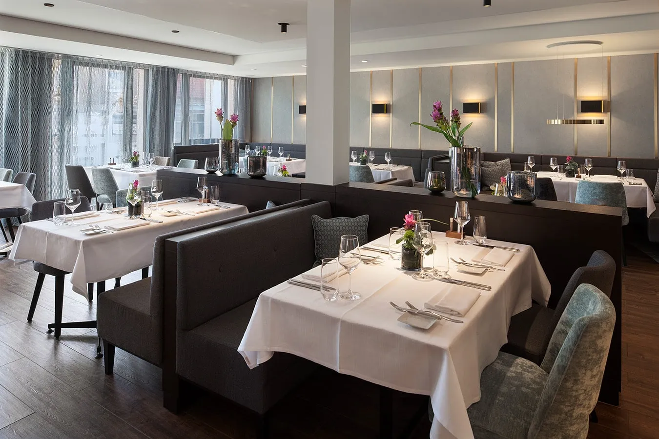 Elegantes Restaurant mit gedeckten Tischen, weißen Tischdecken, Blumenvasen und modernen Paneelen an den Wänden.
