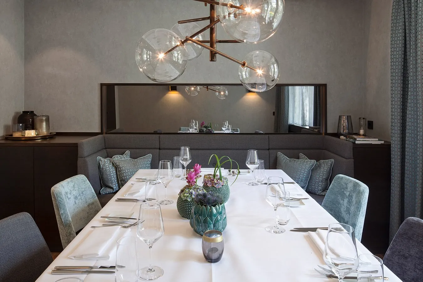Elegant gedeckter Esstisch mit weißen Tischdecken, Blumenarrangement und modernen Glaslampen über einem Sofa in einem stilvollen Raum.