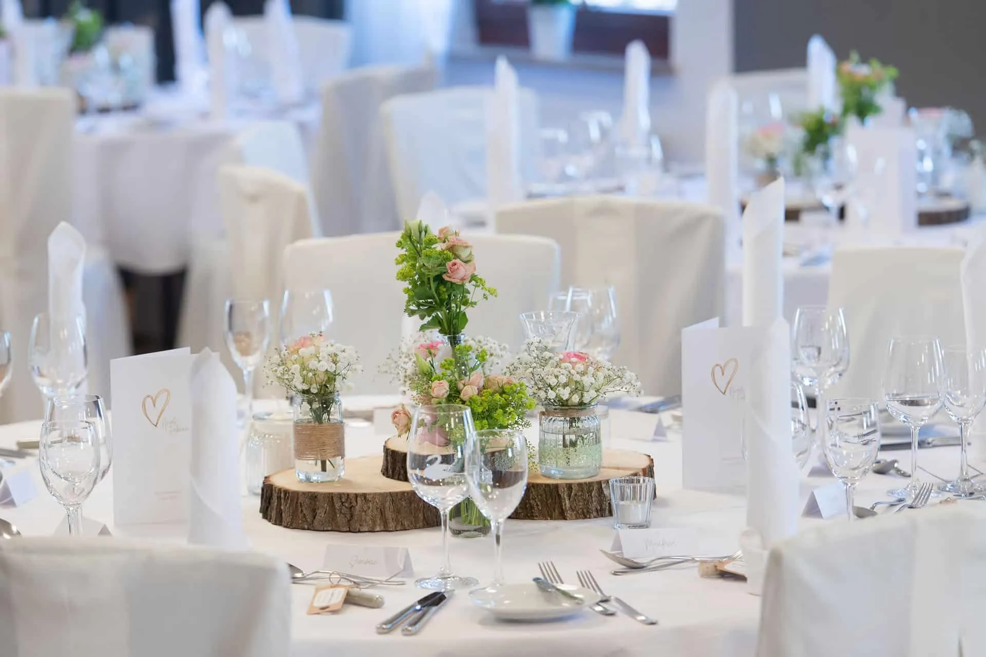 Hochzeitsdekoration: Rund um Tisch, weiße Tischdecken, Blumenarrangements in Vasen auf Baumscheiben, Gläser und Besteck angeordnet.