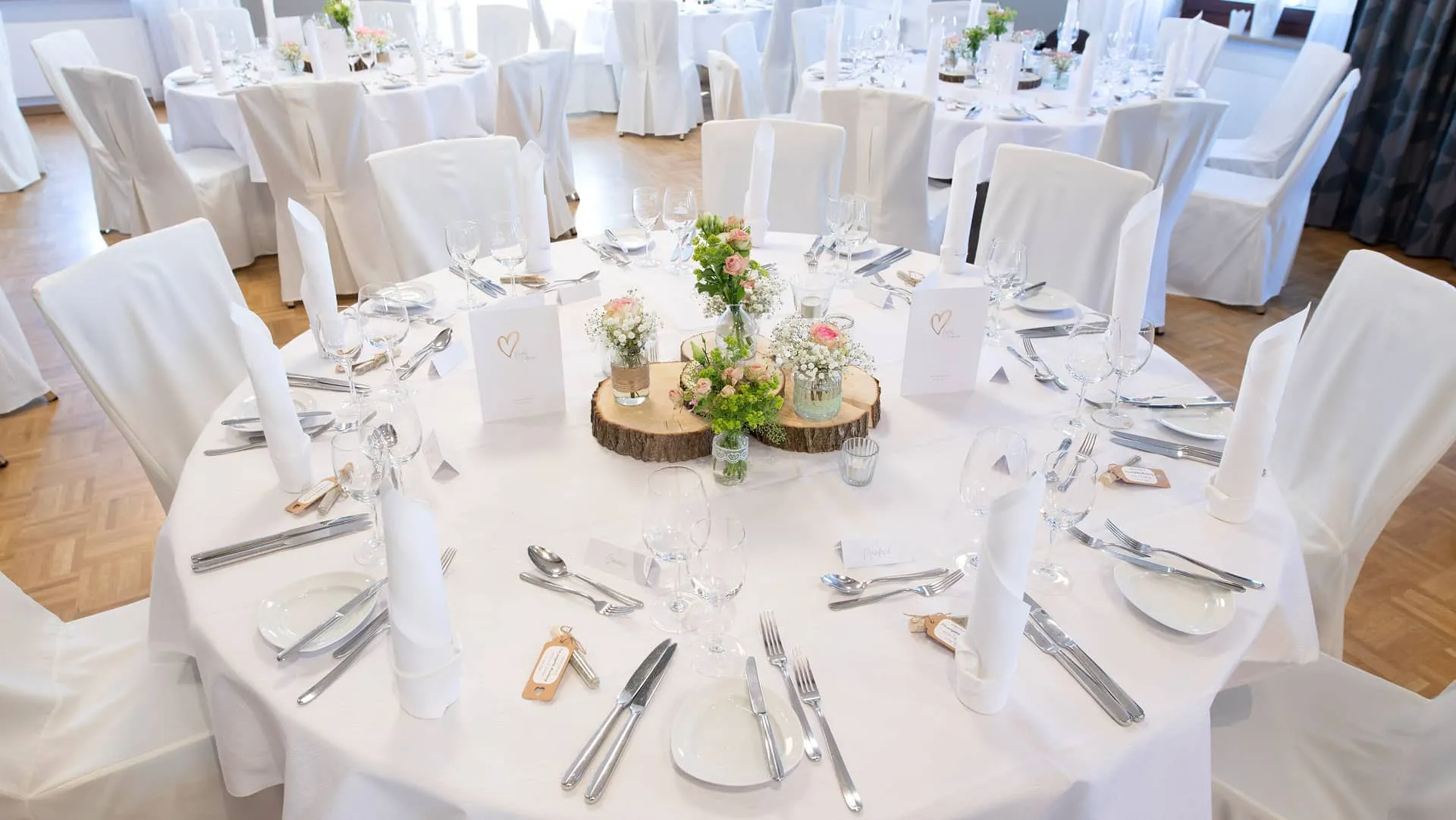 Rund gedeckter Hochzeitstisch mit weißen Hussen, Menükarte, Blumendekoration und Besteck in festlichem Raum.