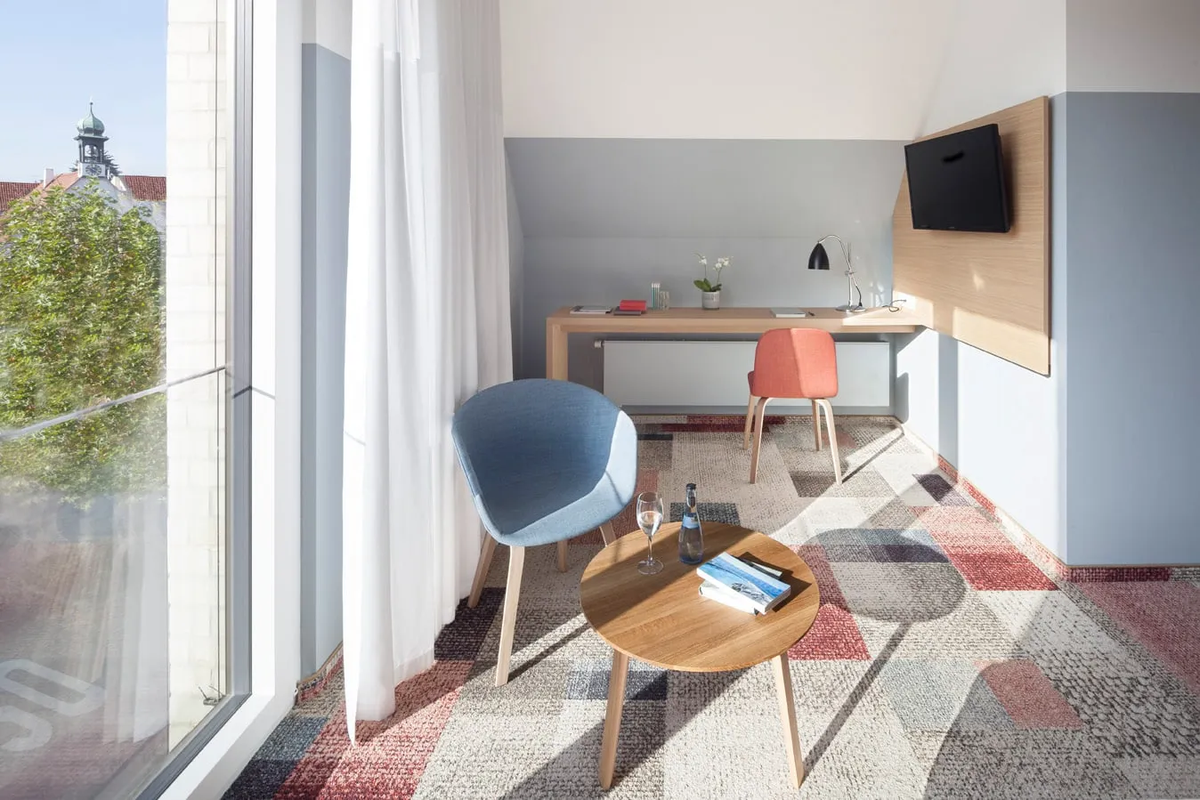 Helles Hotelzimmer mit Schreibtisch, zwei Stühlen, rundem Tisch, Fernseher, Fenster mit Vorhängen und Aussicht auf Stadtgebäude.