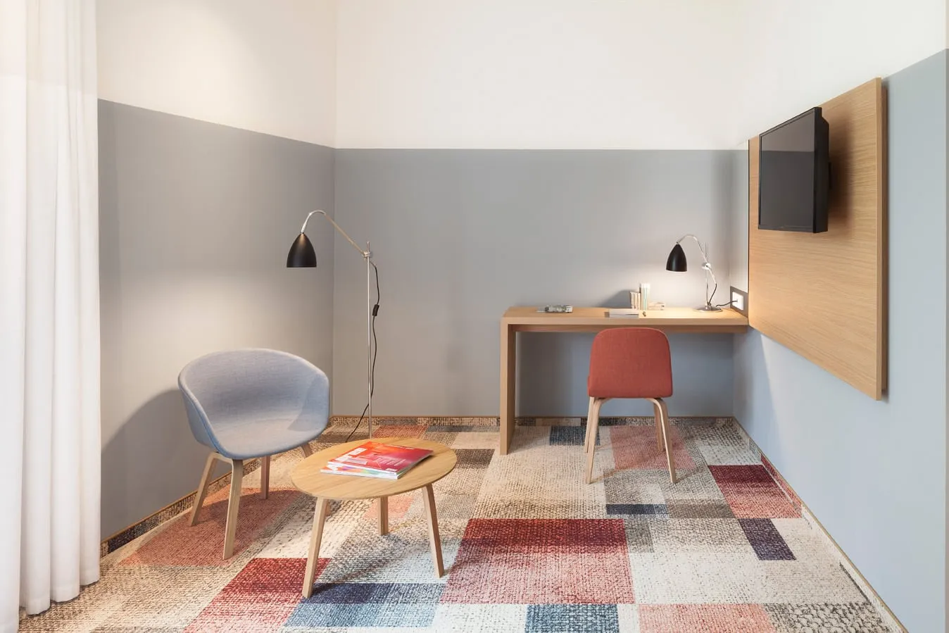 Moderner Raum mit blauem Sessel, Holztisch, rotem Stuhl, Schreibtischlampe und Wandfernseher, auf buntem Teppich.