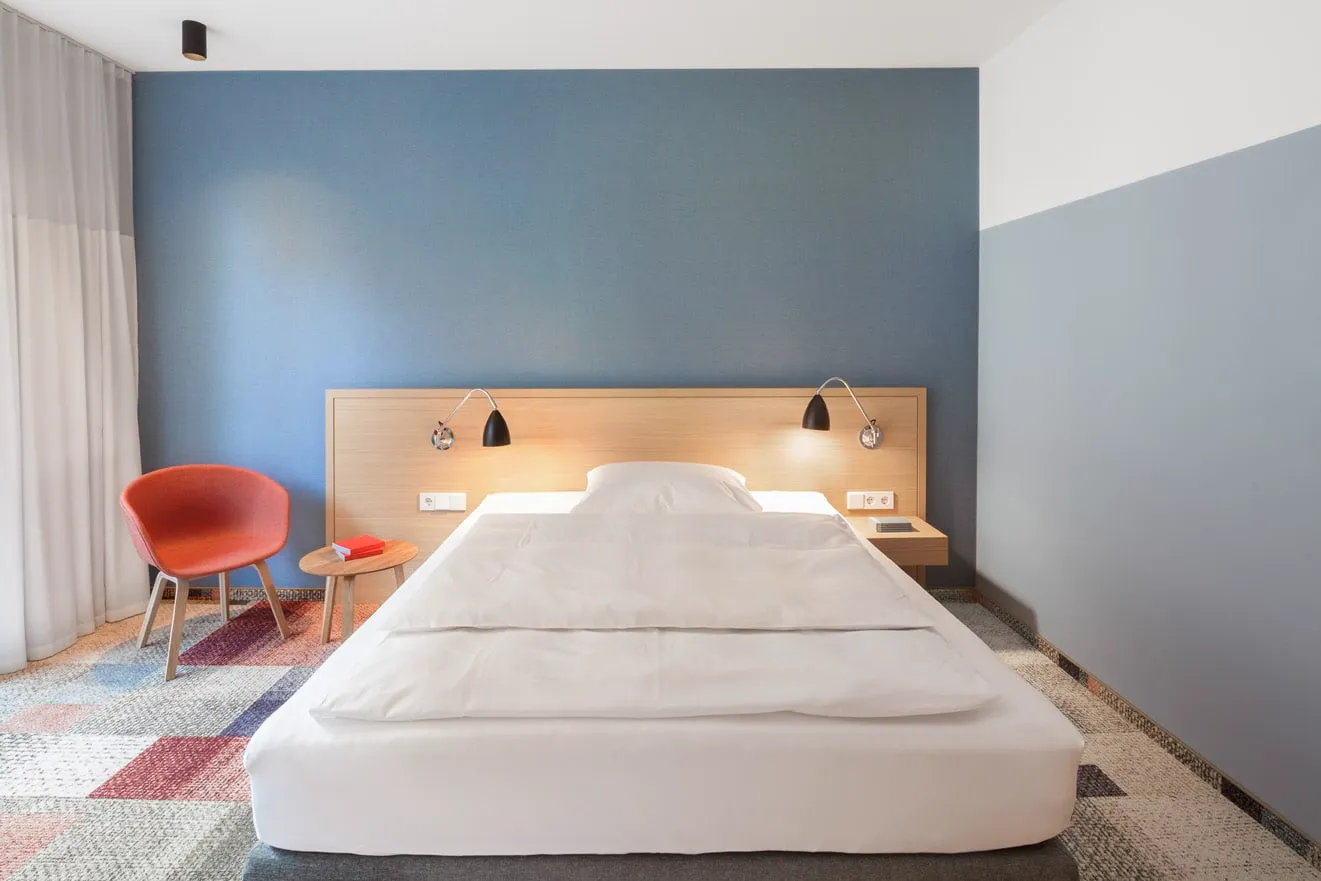 Einzelbett im modernen Hotelzimmer mit blauem Kopfteil, roten Sessel und Nachttisch mit Buch, auf buntem Teppich.