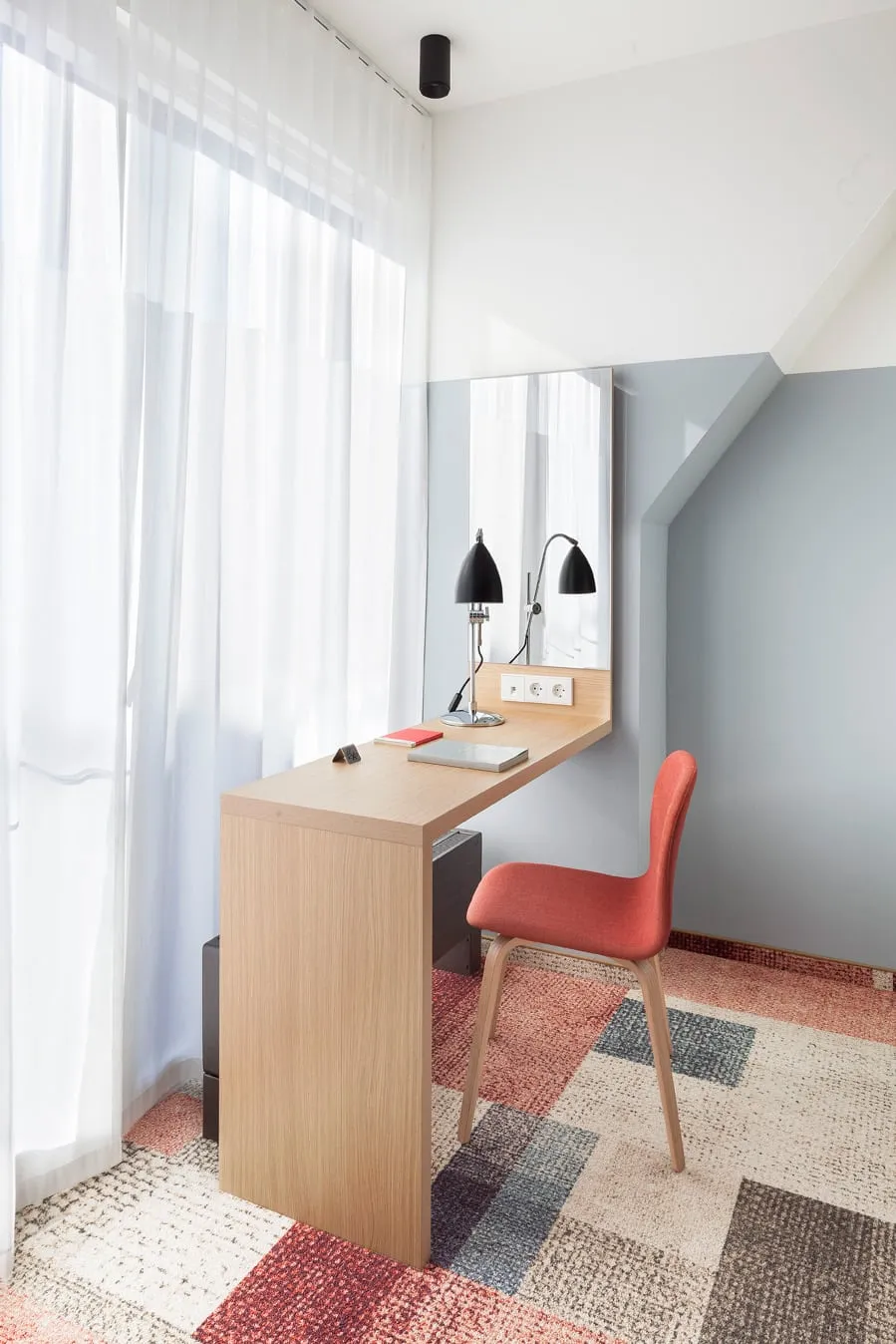 Moderner Arbeitsplatz mit Holzschreibtisch, roten Stuhl, Tischlampe und Laptop vor einer großen Fensterfront mit weißen Vorhängen.