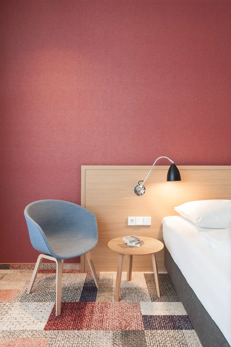 Hotelzimmer mit orangener Wand, blauem Stuhl, Holztisch, Stehlampe und einem Bett auf gemustertem Teppichboden.