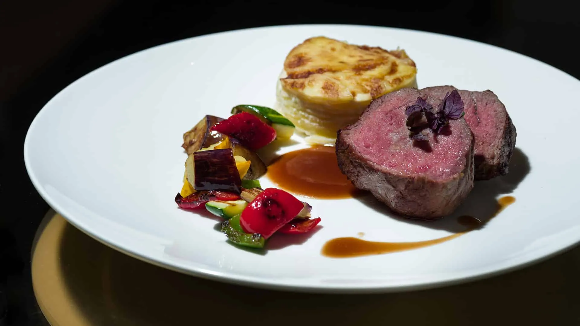 Rosa gebratenes Rinderfilet mit Bratensauce, buntem Gemüse und Kartoffelgratin auf einem weißen Teller.