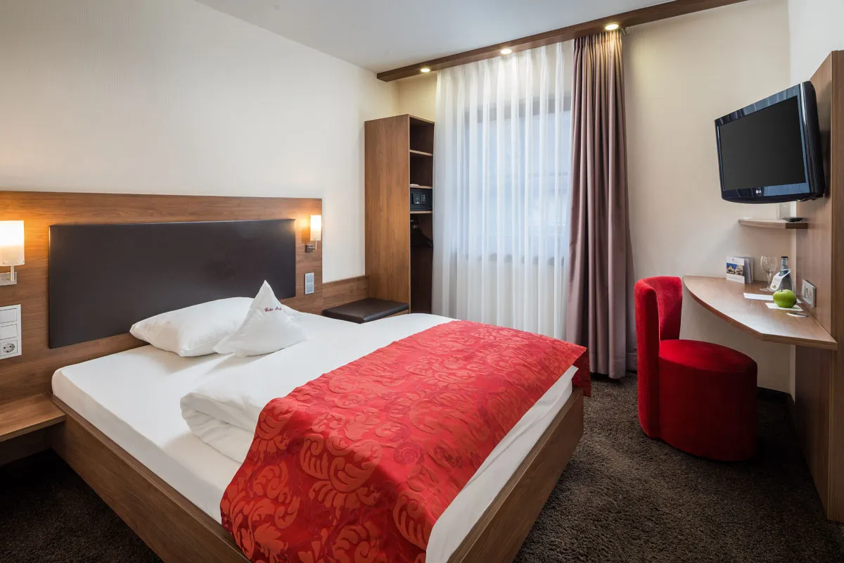 Hotel-Restaurant-Schwanen-Queen-Size-Einzelbett-Zimmer Modernes Hotelzimmer mit Einzelbett, roter Decke, Schreibtisch, rotem Stuhl und Wandfernseher, gedämpfte Beleuchtung.