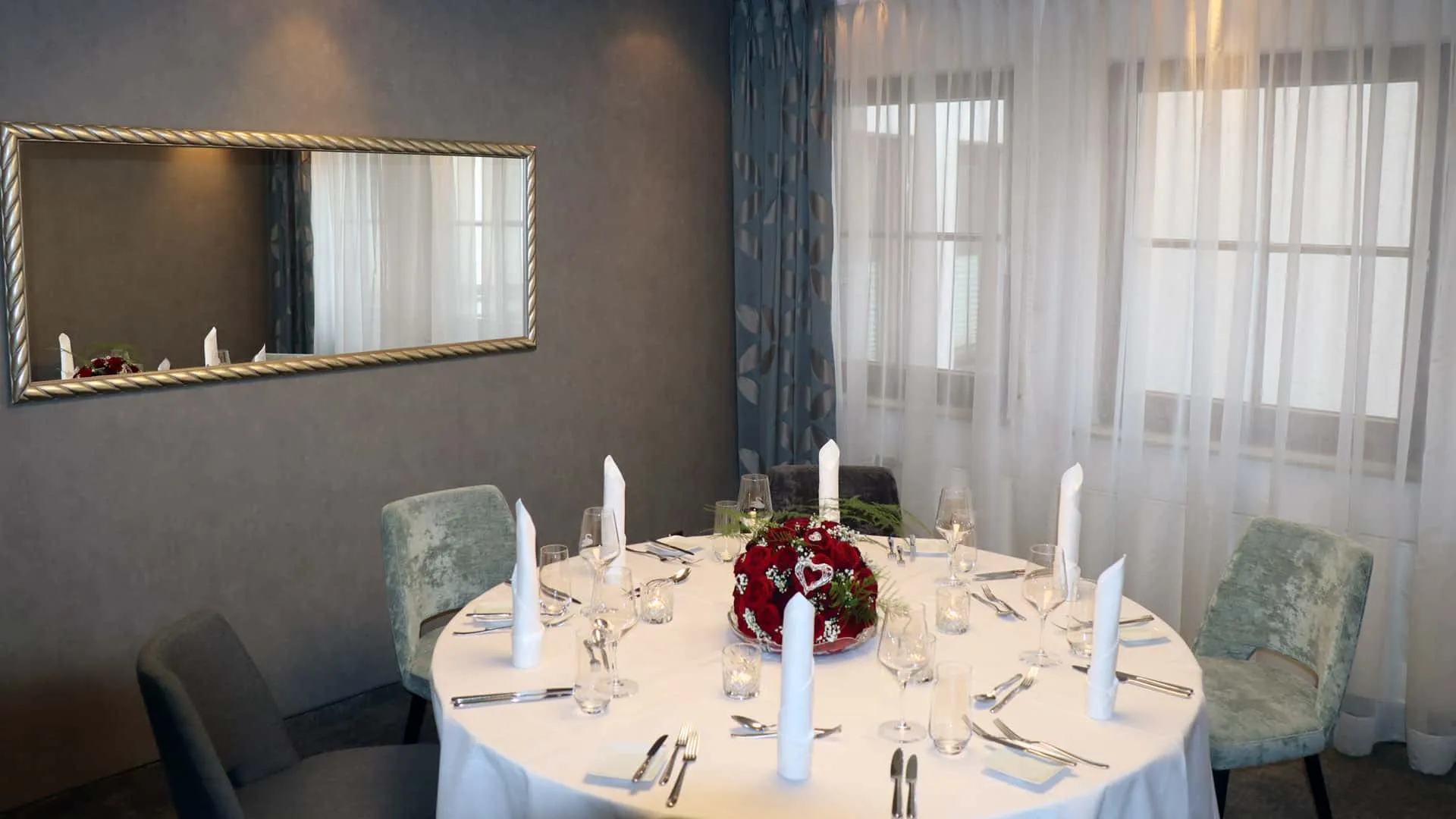 Runder Tisch im Restaurant, gedeckt mit weißen Servietten, Besteck, Gläsern und rotem Blumengesteck, Spiegel an der Wand.
