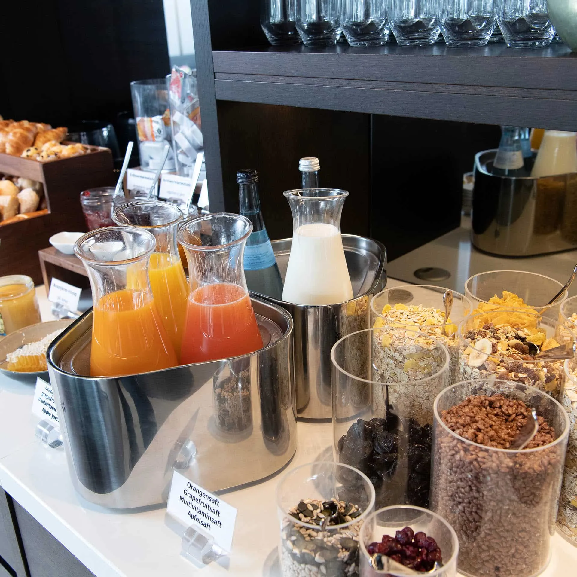 Frühstücksbuffet mit Säften, Milch in Karaffen, verschiedenen Müslisorten und Gebäck auf einem Tisch arrangiert.