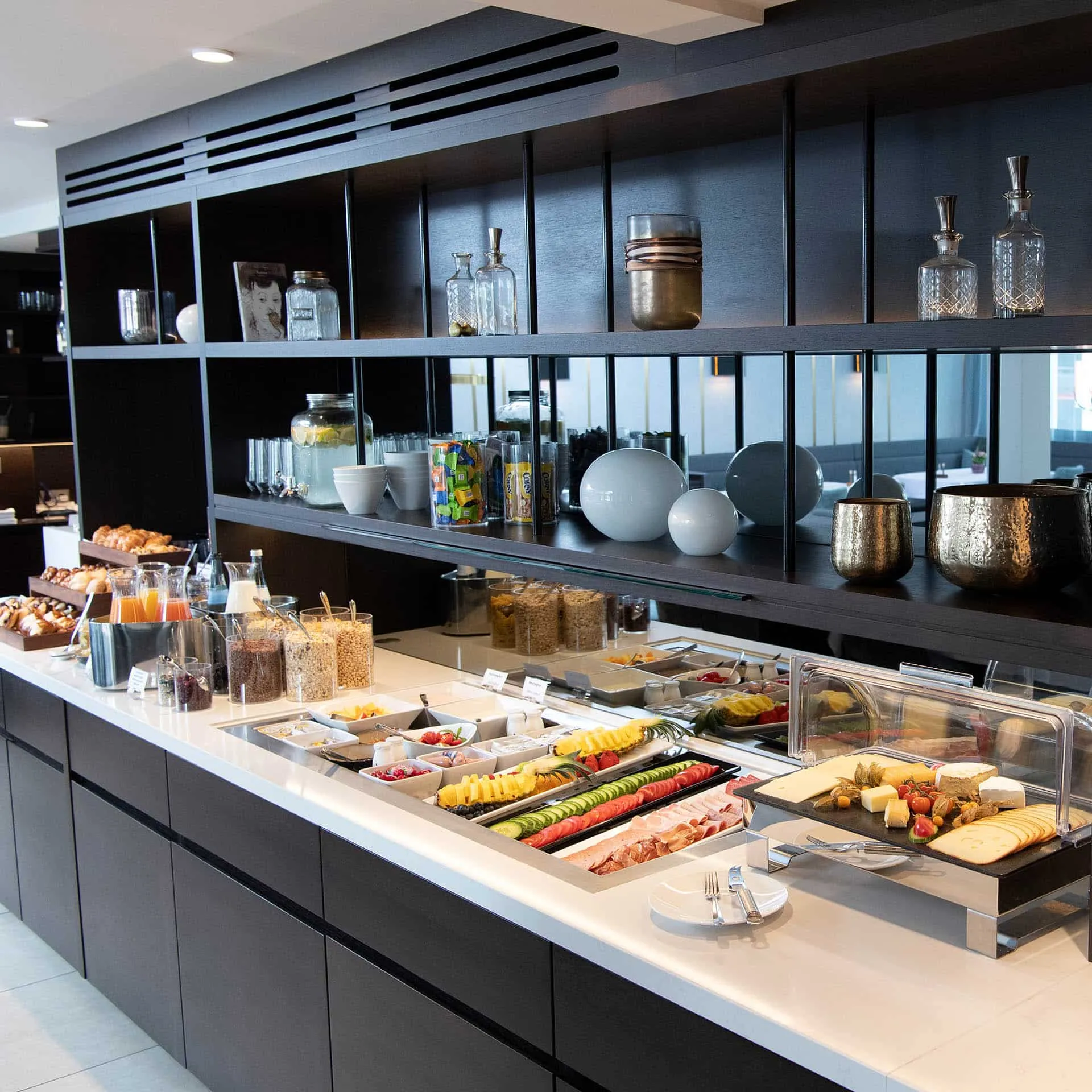 Hotelbuffet mit verschiedenen Frühstücksoptionen, darunter Obst, Brot, Müsli, und Getränke. Elegantes, modernes Design.