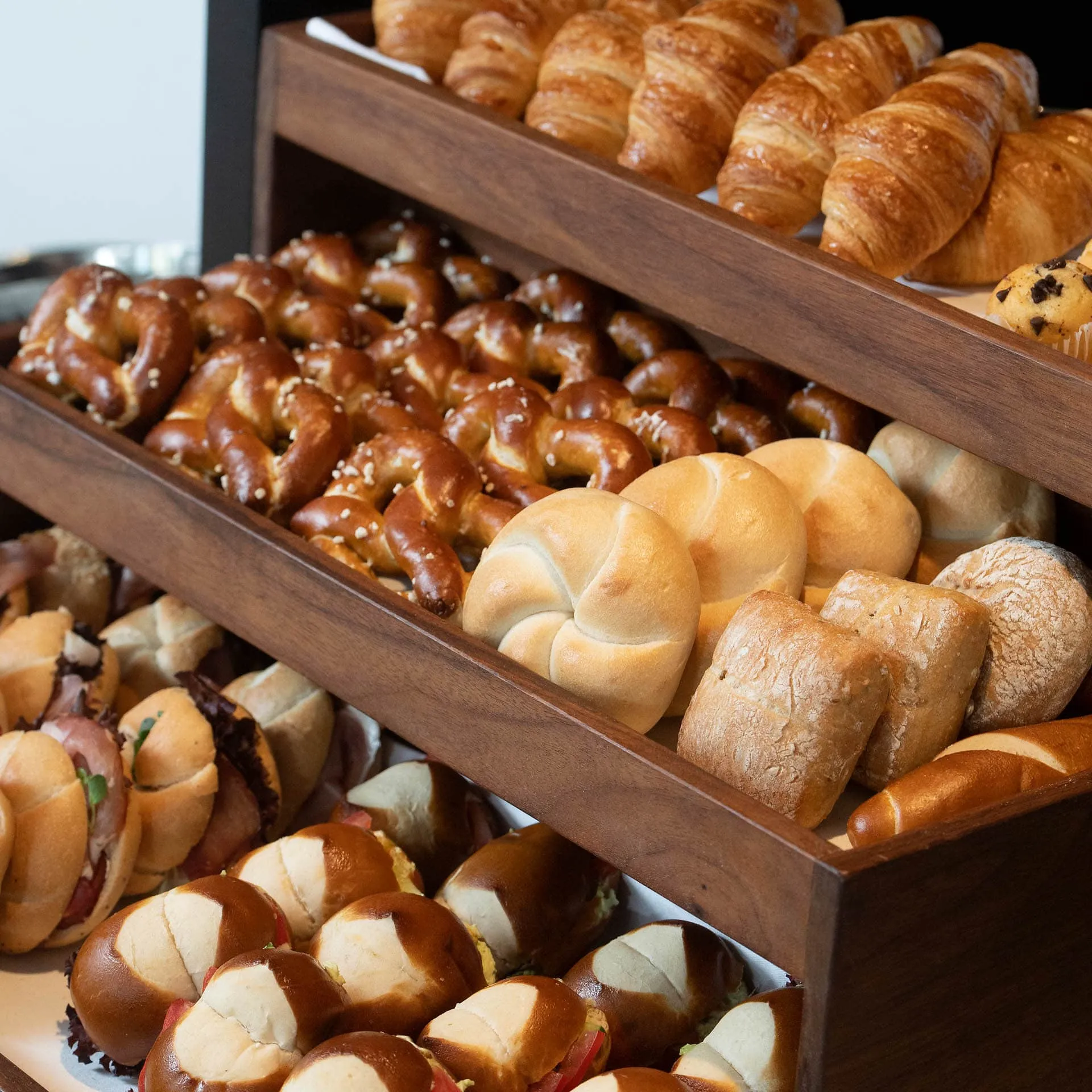 Verschiedene Backwaren auf Holzregalen, darunter Croissants, Brezeln, Brötchen und belegte Laugenbrötchen.
