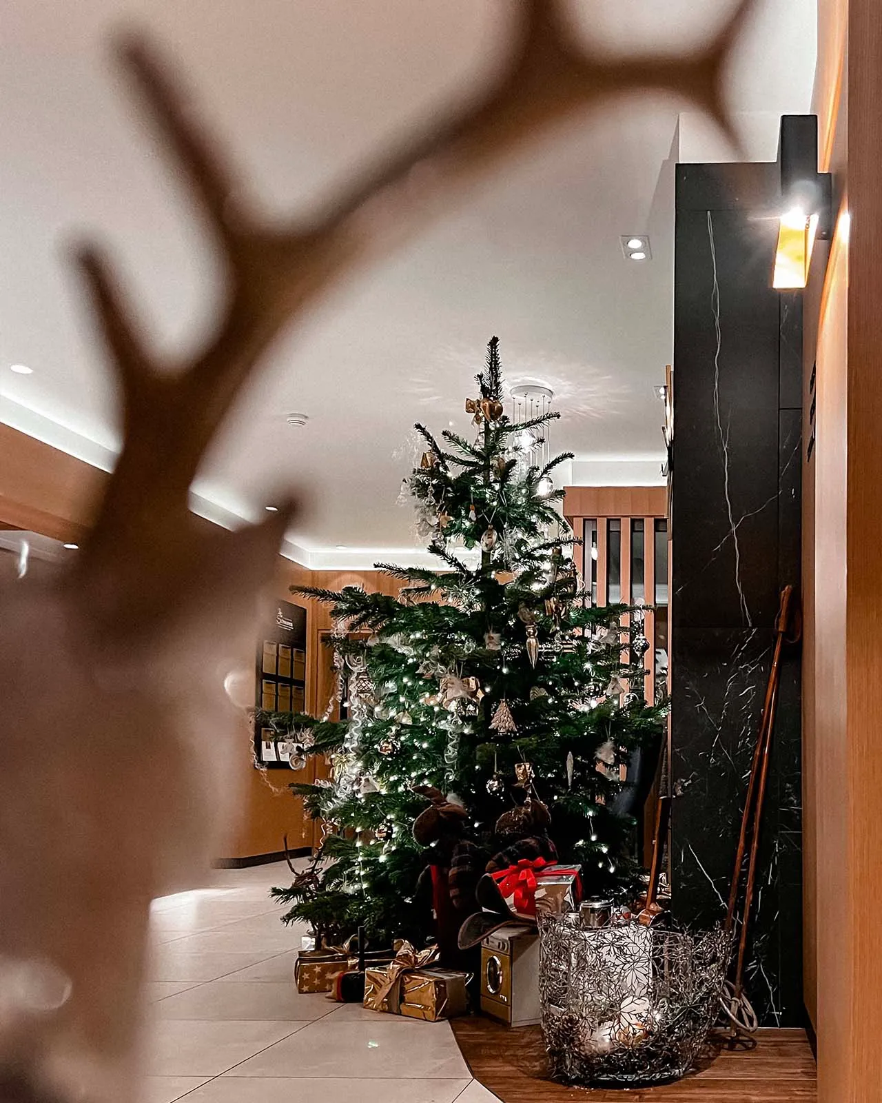 Weihnachtsbaum mit Lichtern und Geschenken in einem modernen Wohnzimmer, unscharfe Rentiergeweihe im Vordergrund.