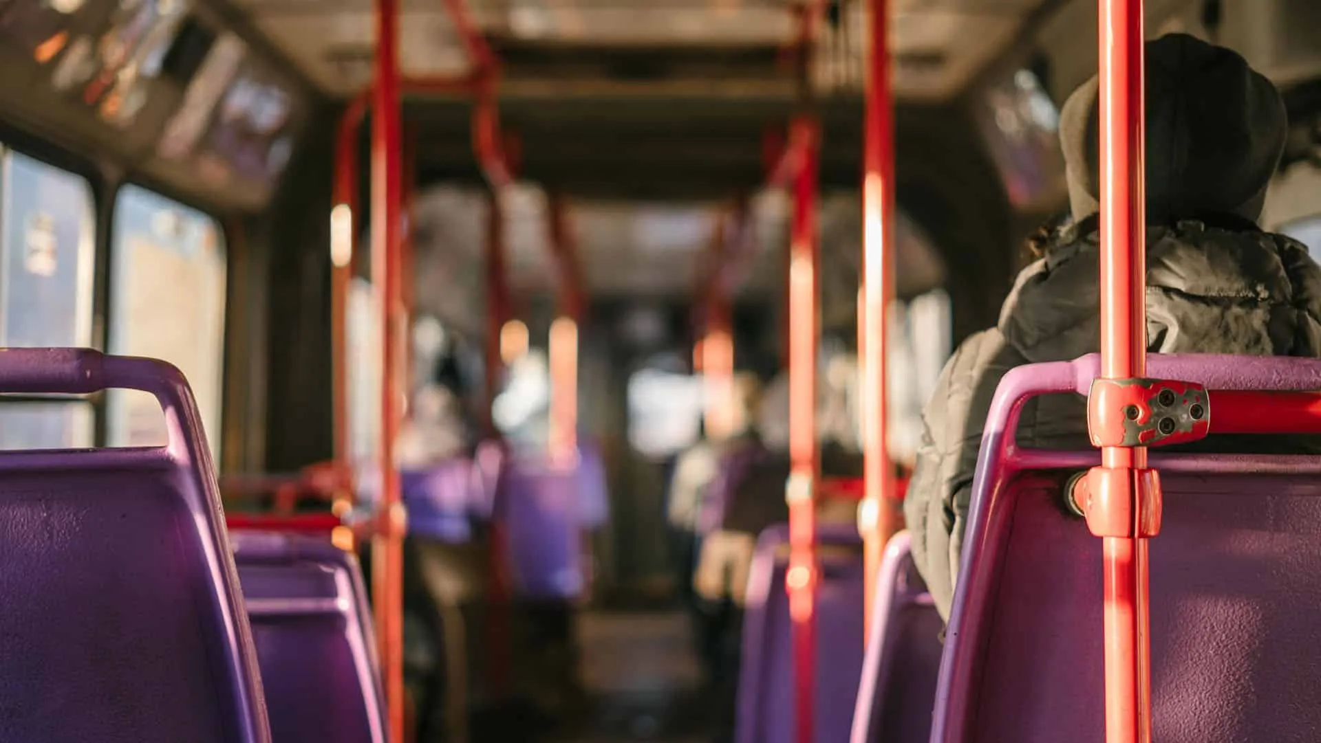 Innenraum eines Busses mit lila Sitzen und roten Haltegriffen, Fahrgäste in Winterkleidung sitzen im Sonnenlicht.