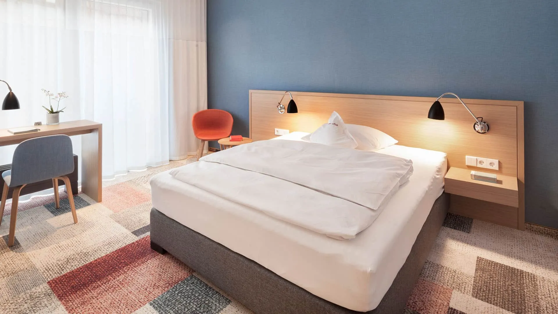 Moderner Hotelraum mit Bett, Schreibtisch und Stuhl. Blaue Wand, rote Akzentfarbe, Teppichmuster in Beige- und Rottönen.