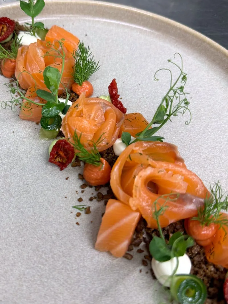 Gericht mit Lachsrosen, Erbsensprossen, Dill, Creme und Kaviar auf beigefarbenem Teller kunstvoll angerichtet.