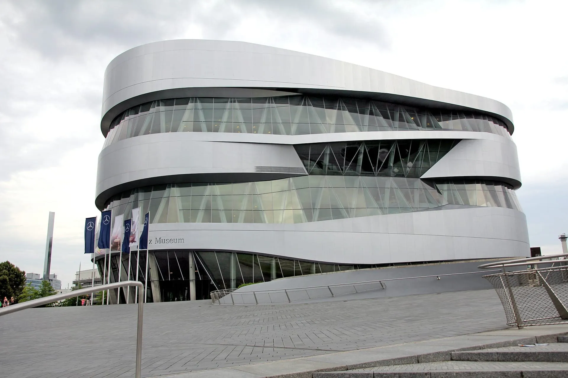 imgi_12_Hotel-Schwanen-Arrangement-Stuttgart-Daimler-Museum Modernes Gebäude mit geschwungener Glas- und Stahlfassade, Fahnen im Vordergrund. Bewölkter Himmel darüber.
