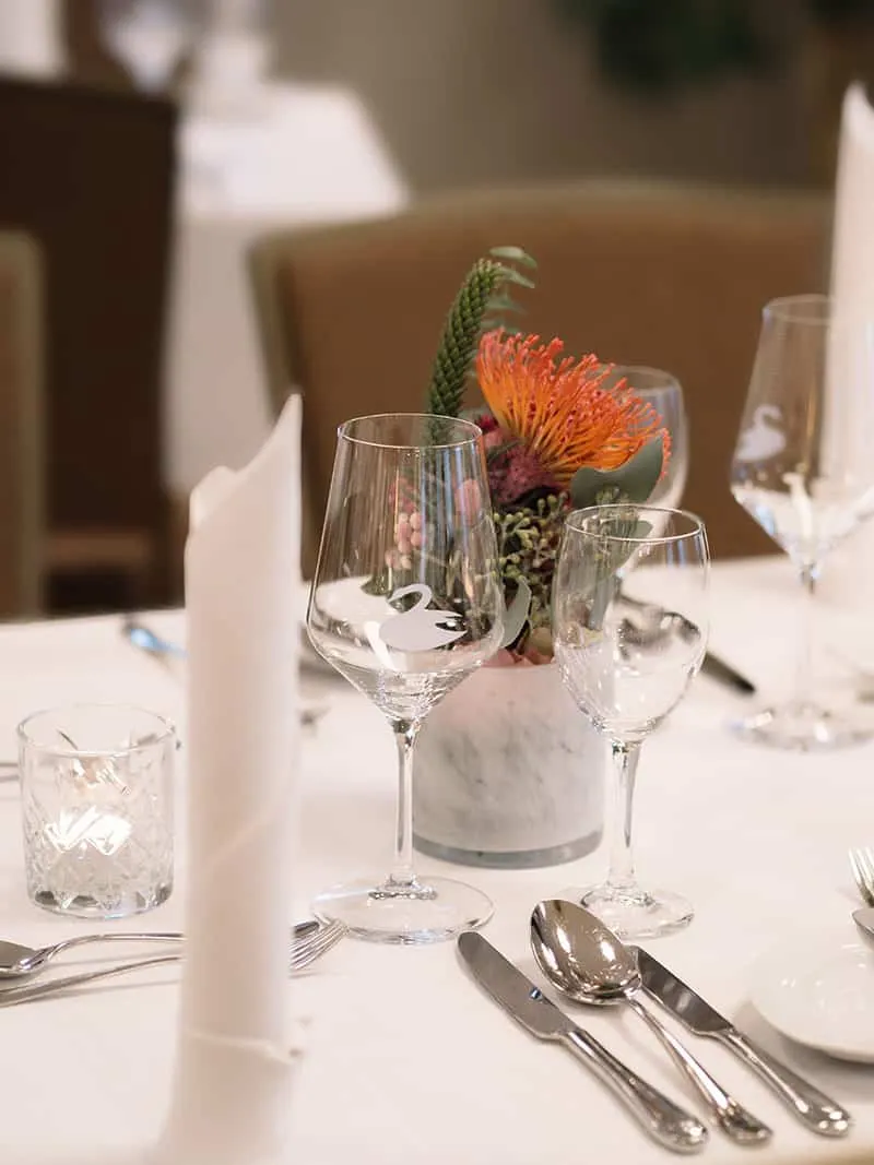 Eleganter, gedeckter Tisch mit Weingläsern, Besteck, weißen Servietten und einer Blumenarrangement in einem Marmorgefäß.