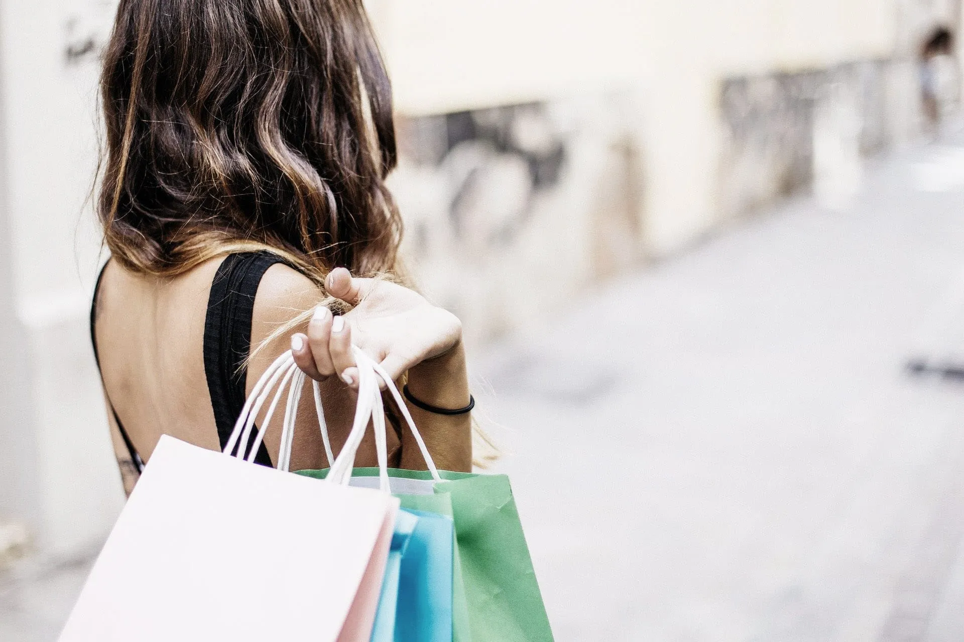 imgi_13_Hotel-Schwanen-Arrangement-Shopping Frau mit Einkaufstüten geht durch eine Stadtstraße, Rücken zur Kamera, pastellfarbene Tüten in der Hand.