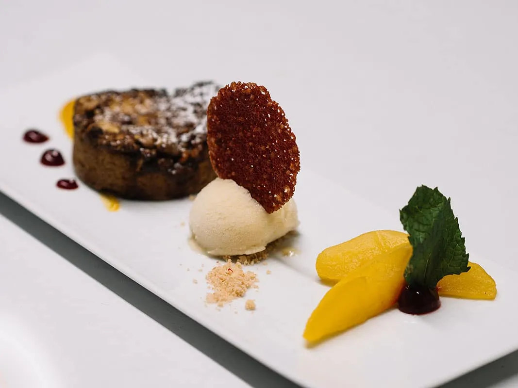 Dessert auf Teller: Schokoladenkuchen, Vanilleeis, Mangospalten, Minzblatt und Fruchtsauce, garnieret mit knusprigem Keks.