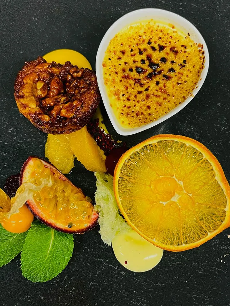 Dessertplatte mit Crème Brûlée, Walnusskuchen, Passionsfrucht, Orangenscheibe, Minzblättern und Fruchtsoßen auf schwarzem Teller.