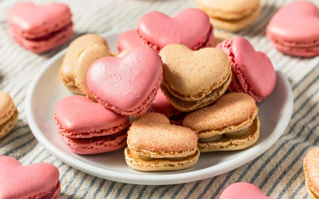 Herzförmige rosa und beige Macarons auf einem weißen Teller, auf gestreiftem Tuch arrangiert.