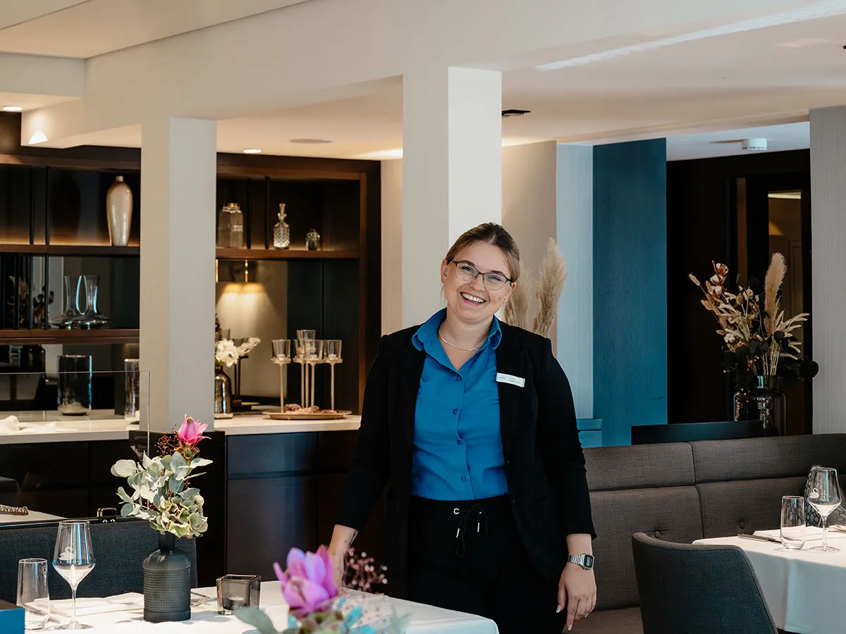 Eine lächelnde Person in Uniform steht in einem Restaurant, umgeben von Blumen und gedeckten Tischen.