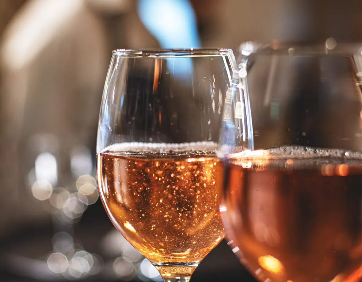 Zwei mit roséfarbenem Sekt gefüllte Weingläser, Schaum und Lichtreflexionen, unscharfer Hintergrund.