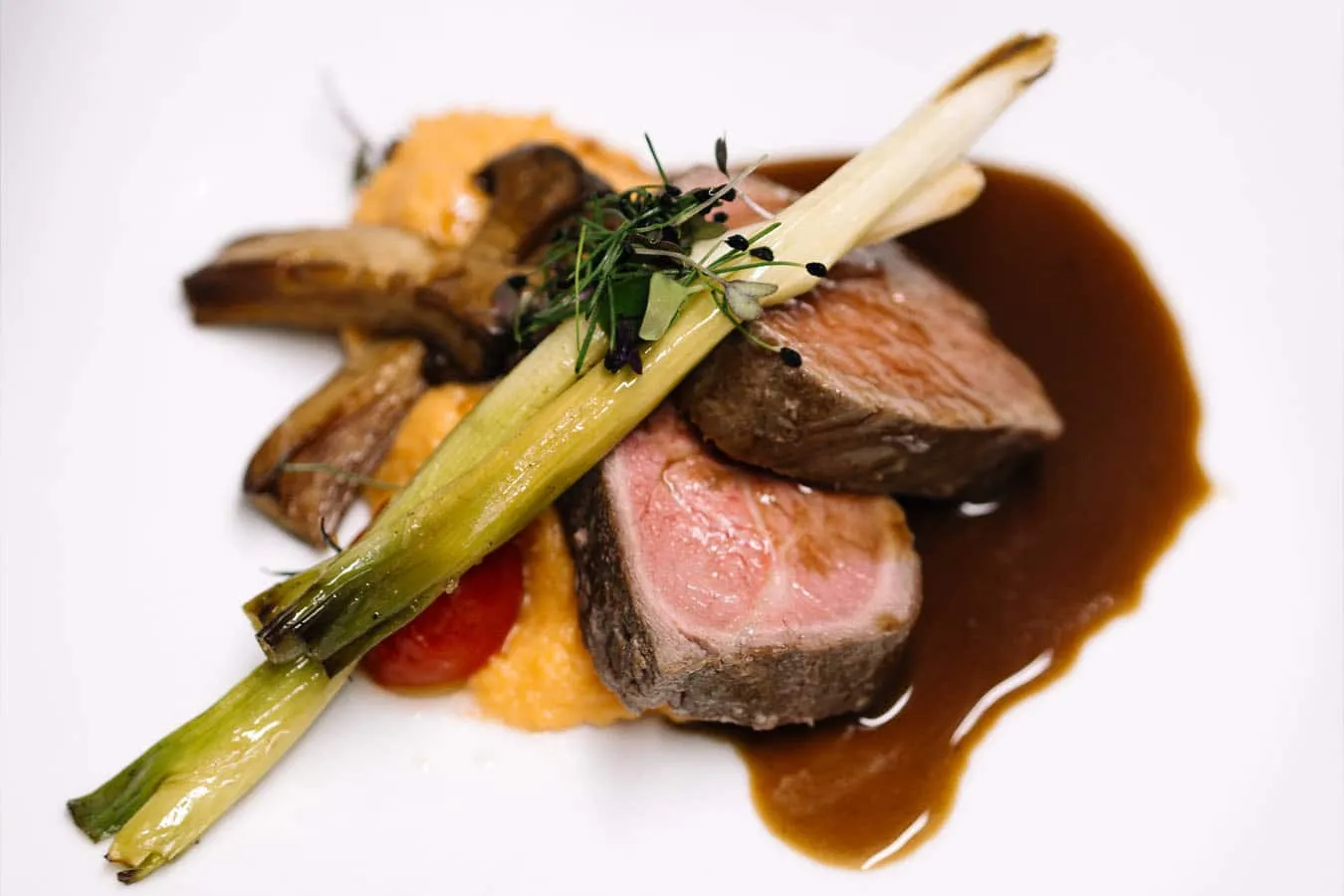 Gebratene Fleischscheiben mit Lauch, Pilzen und Sauce auf cremigem Püree, garniert mit Kräutern auf weißem Teller.