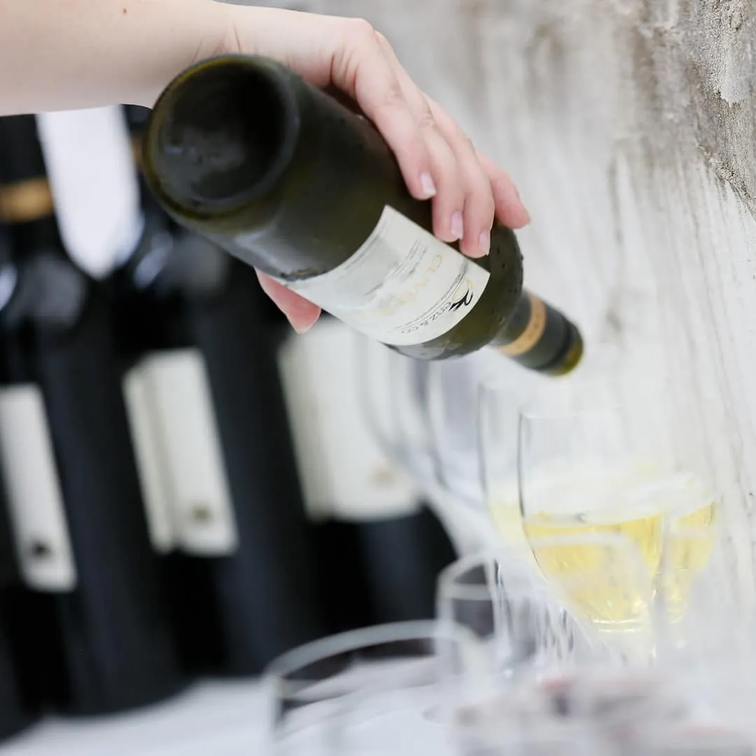 Eine Hand schenkt Weißwein in ein Glas ein; Flaschen stehen im Hintergrund.